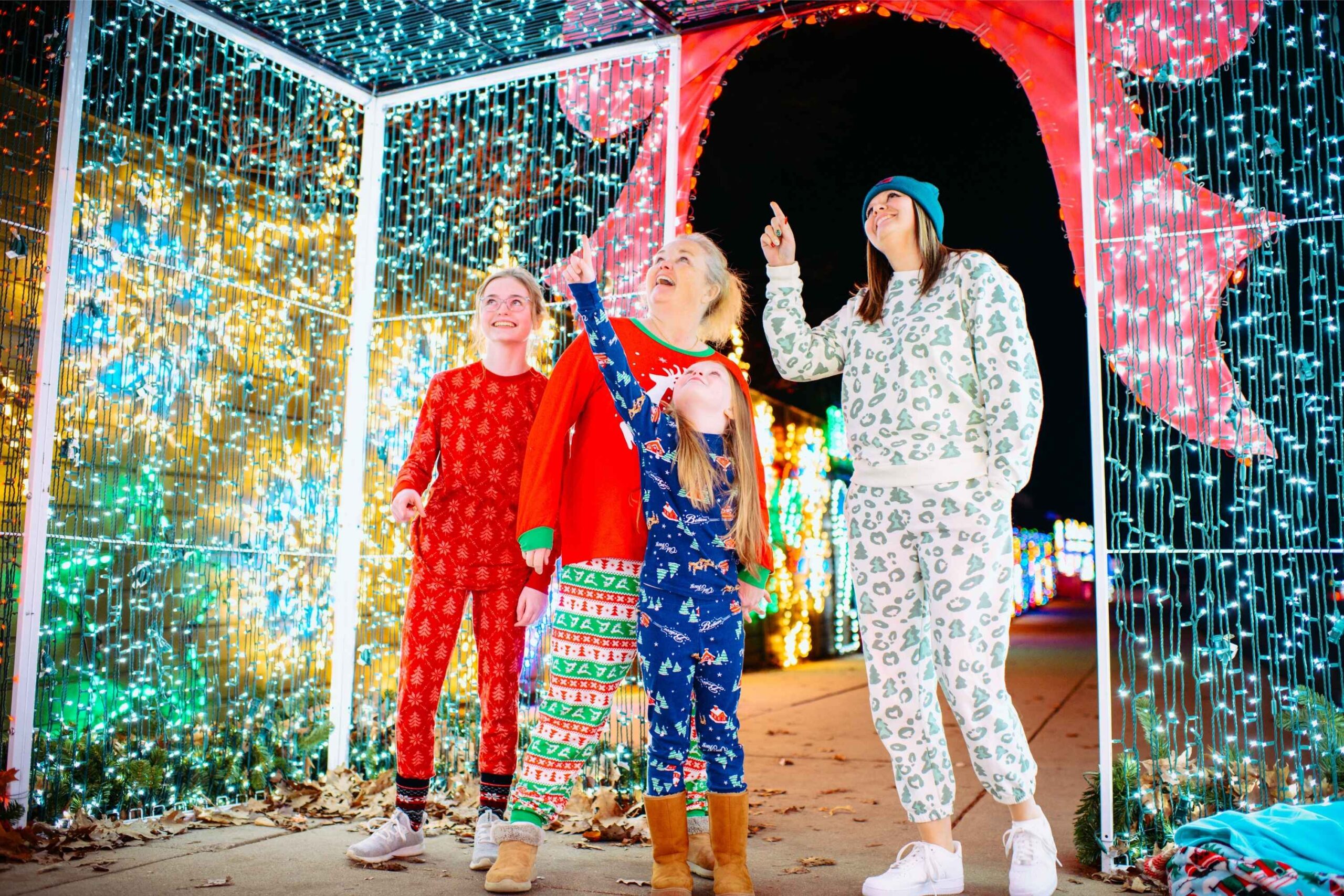 A family walks through a walk-through light tunnel at the Magic of Lights at Pine Knob Music Theatre, pointing at colorful holiday displays.