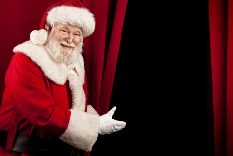 Santa smiles and gestures toward a stage curtain promoting the Miracle on 34th Street show at the Fox Theatre supporting Make-A-Wish Michigan