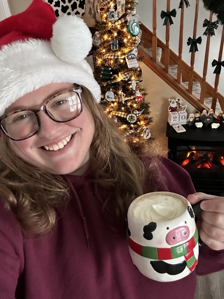 Samantha Whitehead smiling in front of her decorated Christmas tree while holding a mug of rich, dairy-heavy holiday hot cocoa topped with whipped cream.
