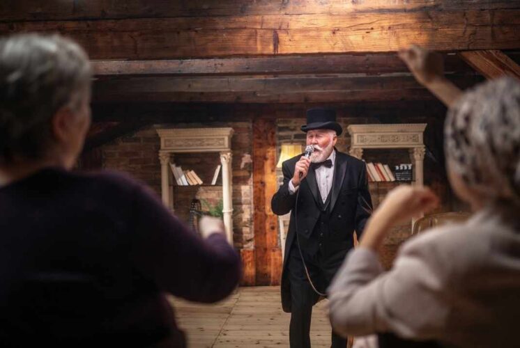 Performer dressed as Scrooge sings onstage at The Hawk Theatre as audience watches during a family friendly holiday show
