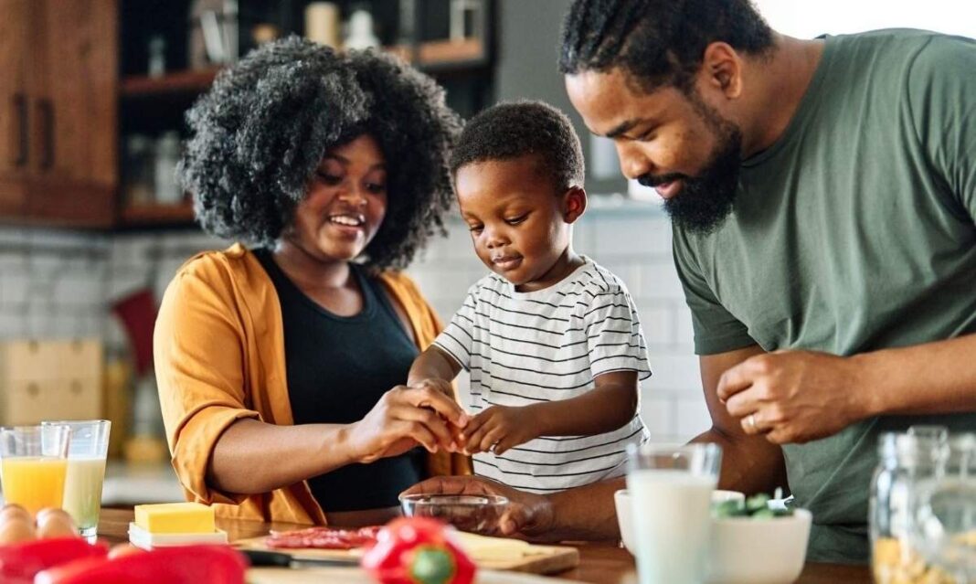 Black family cooking together in a kitchen, helping a young child prepare a healthy meal with milk and vegetables on the counter