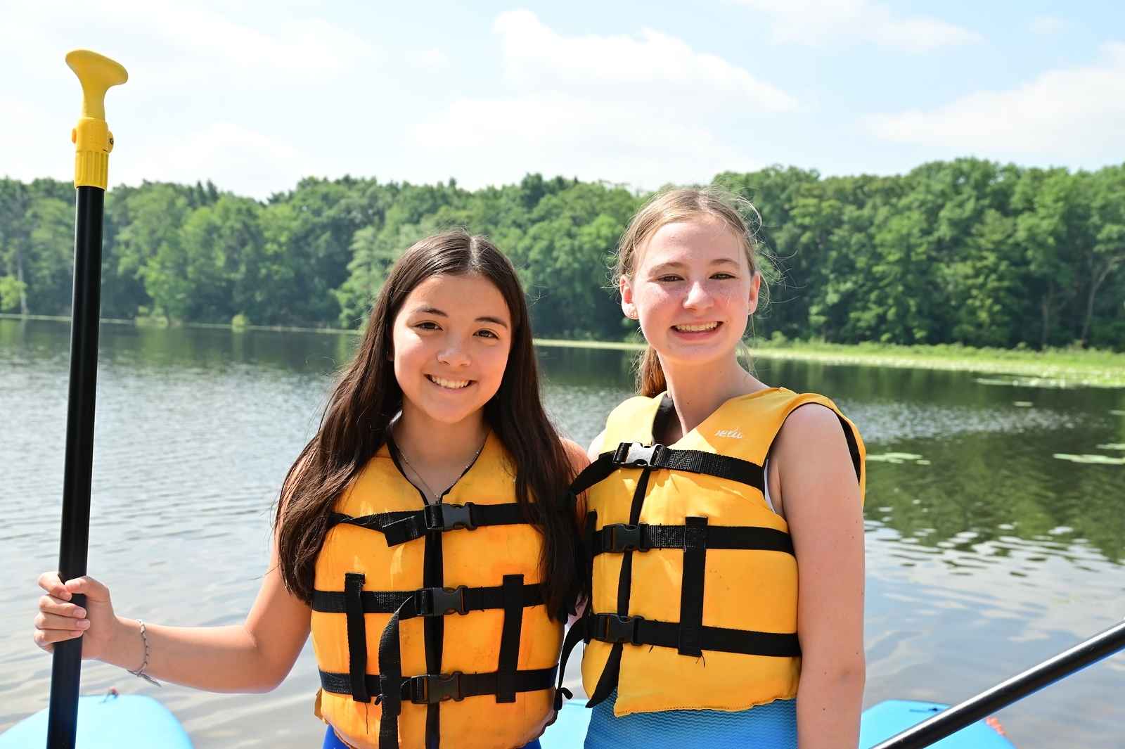 Two campers wearing life jackets smile while paddleboarding on the lake at YMCA Camp Ohiyesa in Holly Michigan.