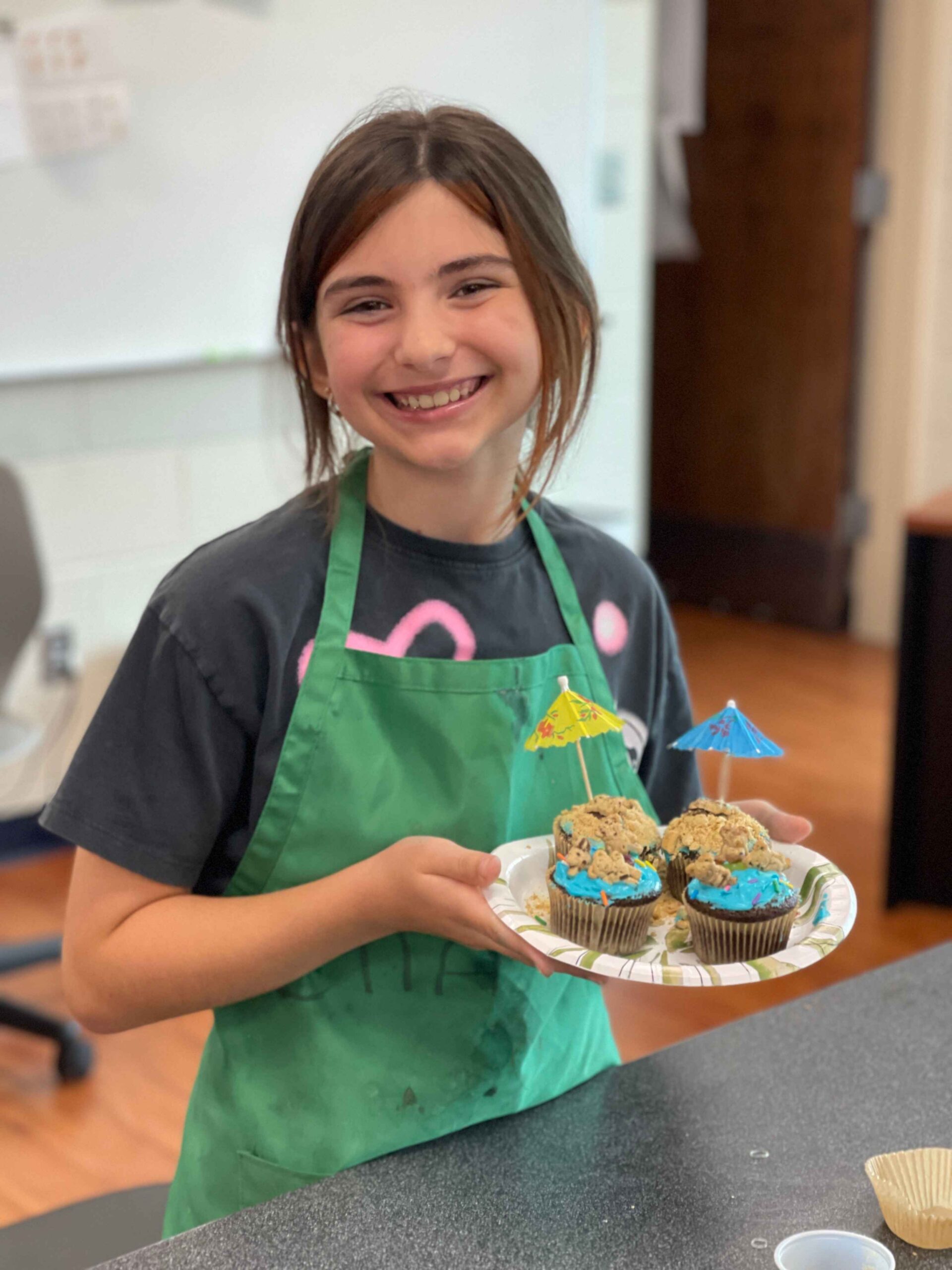 Child smiles while holding decorated cupcakes during a cooking class at Detroit Country Day Camps, showing hands on learning and creativity