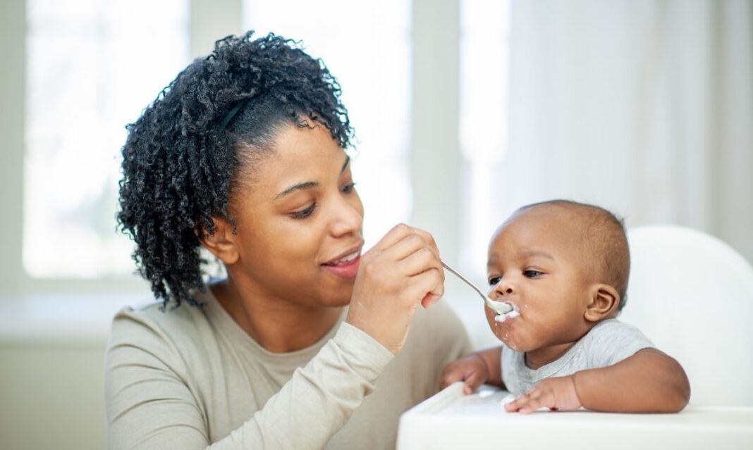 Mom feeds baby pureed food with a spoon in a high chair illustrating how to introduce solid foods safely at 4 to 6 months.
