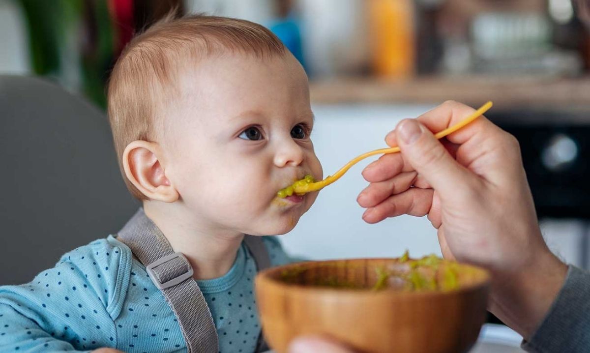 Parent feeds baby pureed vegetables in a high chair demonstrating how to introduce solid foods safely around 4 to 6 months.